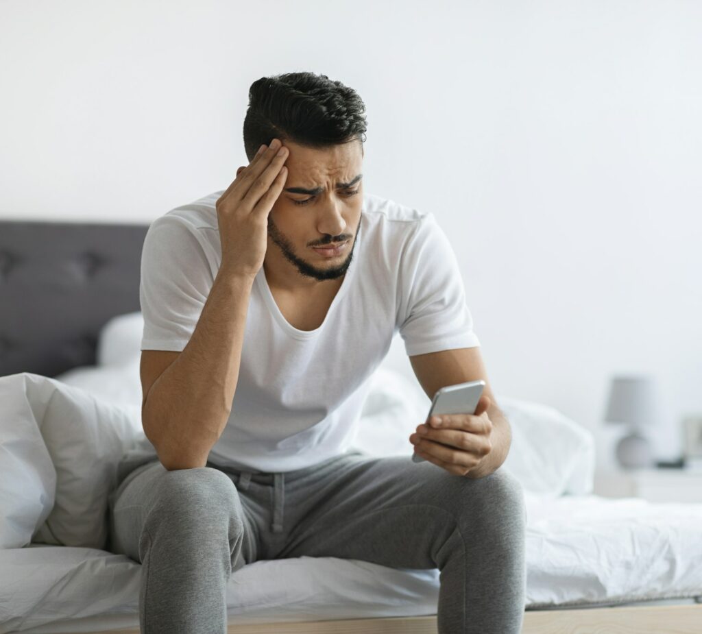 Bad News. Upset Arab Man Sitting On Bed And Looking At Smartphone